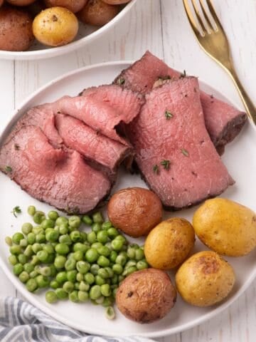 Thinly shaved slices of bottom round roast on a white dinner plate with peas and baby potatoes.