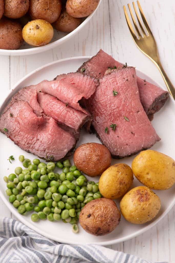 Thinly shaved slices of bottom round roast on a white dinner plate with peas and baby potatoes.