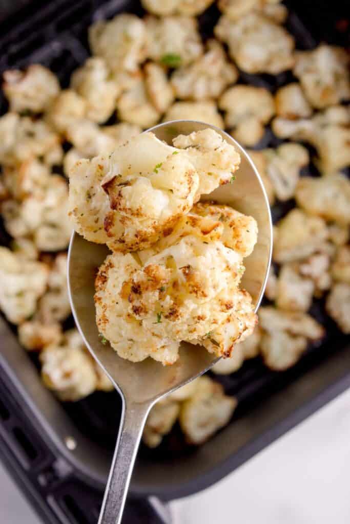 A serving spoon lifting a serving of cauliflower out of the air fryer basket.
