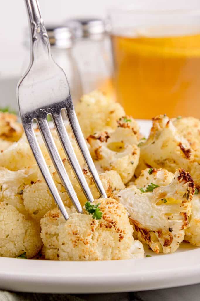 A fork picking up a piece of cauliflower off a white dinner plate.