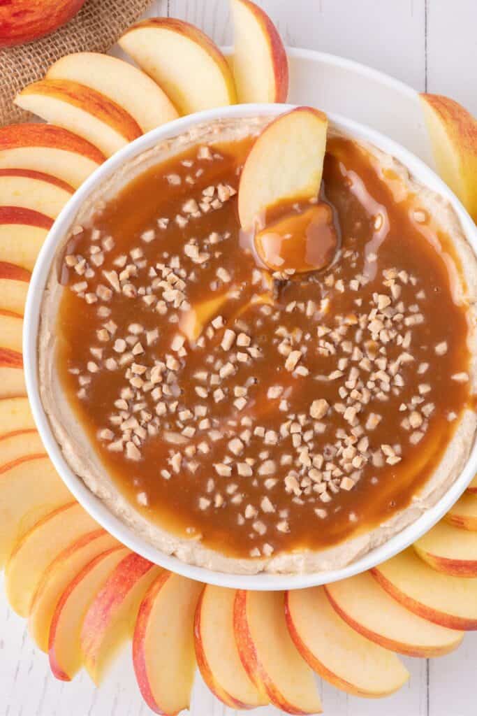 Caramel dip in a bowl surrounded by apple slices. An apple slice is scooping up a bite of caramel dip.