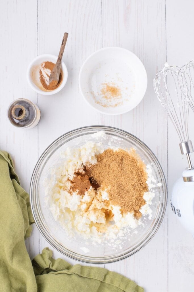 Whipped cream cheese in a glass bowl with brown sugar, vanilla, and cinnamon. A hand mixer is to the right of the bowl.