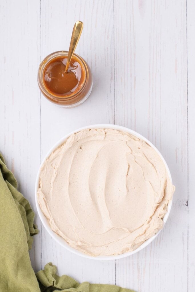 The whipped cream cheese mixture is spooned and smoothed into a bowl. A jar of caramel sauce is sitting next to the dip.