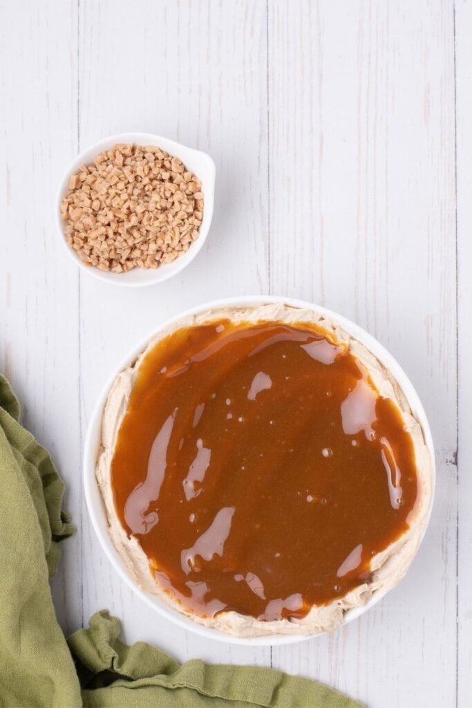 The cream cheese layer has been covered in caramel sauce. A small bowl of toffee bits are right next to the dip.