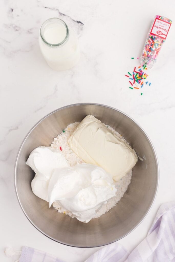 A mixing bowl with Funfetti cake mix, whipped cream, and cream cheese.