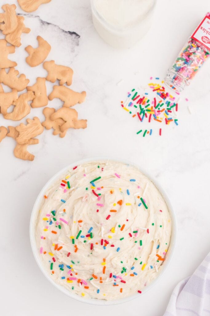 Completed Funfetti dip in a white bowl. Top of dip is decorated with rainbow sprinkles. A pile of animal crackers, jar of rainbow sprinkles, and a glass of milk surround the bowl.