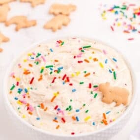 Completed Funfetti dip in a white bowl. Dip is decorated with rainbow sprinkles and a single animal cracker.