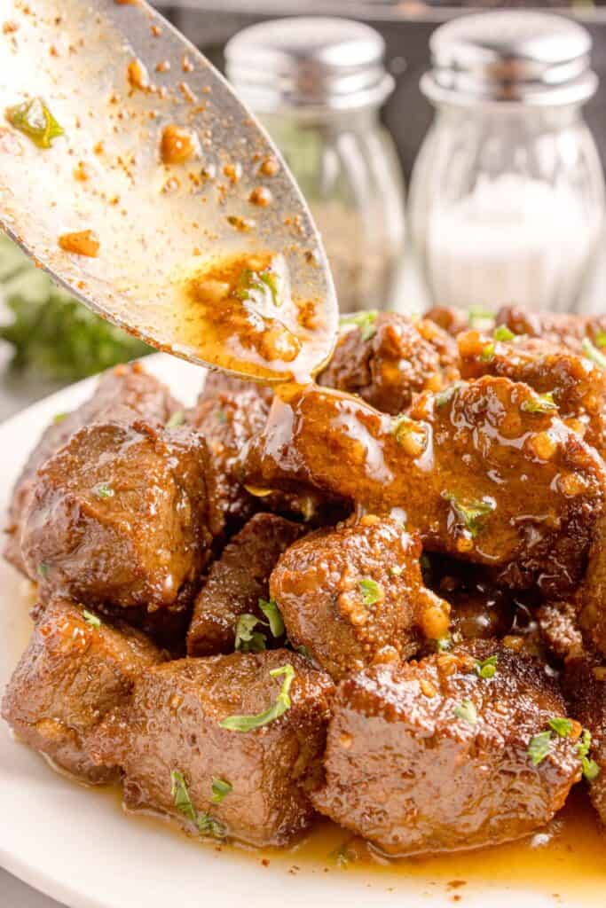 Steak bites on a white dinner plate. A serving spoon is pouring garlic butter sauce over the top of the bites.