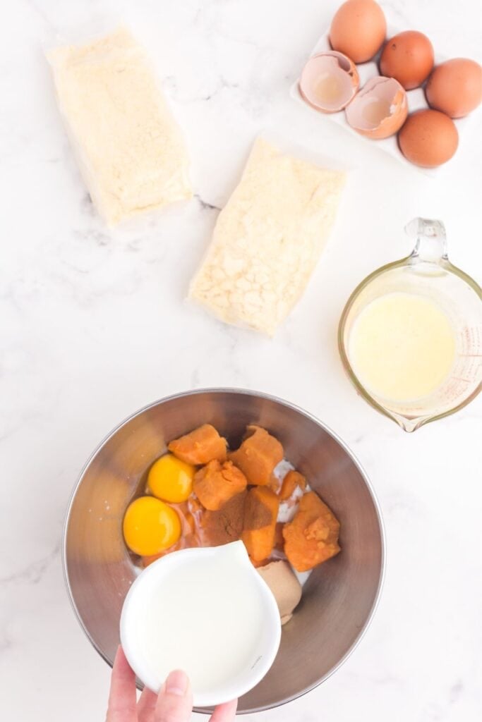 Milk being poured into a mixing bowl of eggs, brown sugar, cinnamon, and cooked sweet potatoes (yam).