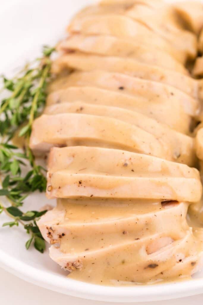 Sliced turkey breast smothered in turkey gravy on a white serving plate with a fresh thyme garnish.