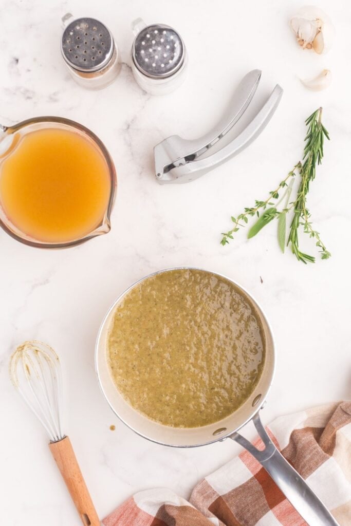 Melted butter and spices in a saucepan. Saucepan is surrounded by fresh herbs, garlic, a garlic crusher, salt and pepper shakers, and a measuring cup of turkey broth.