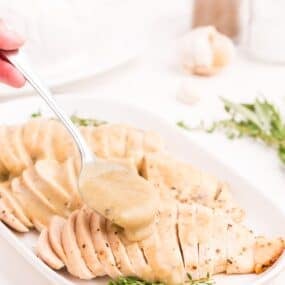 White serving dish with three sliced turkey breasts. A spoon is pouring gravy over the top of the turkey breasts.