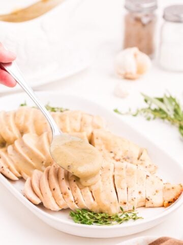 White serving dish with three sliced turkey breasts. A spoon is pouring gravy over the top of the turkey breasts.