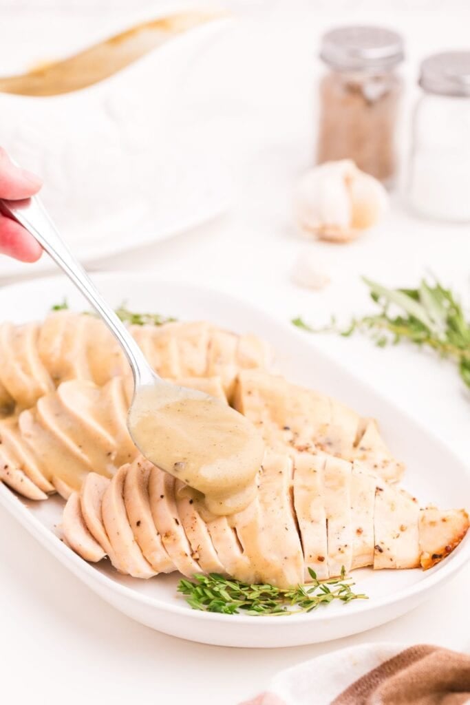 White serving dish with three sliced turkey breasts. A spoon is pouring gravy over the top of the turkey breasts.