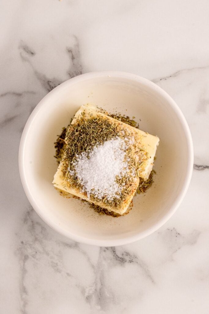 A white bowl with butter, salt, rosemary, thyme, sage, garlic powder, smoked paprika, and pepper.