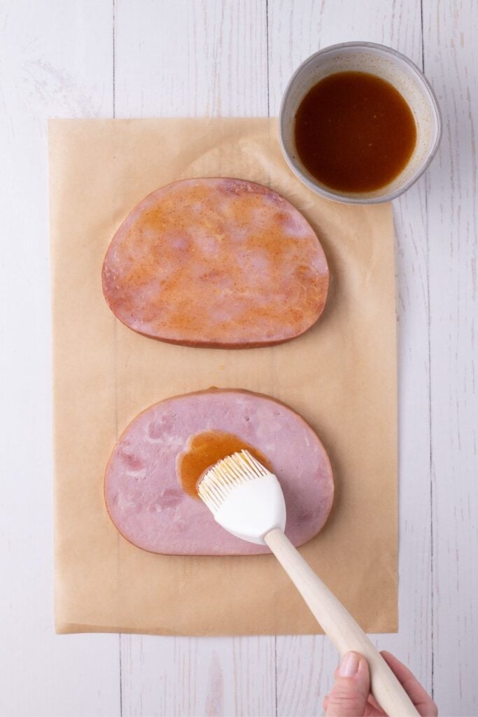 Two ham steaks are placed on a piece of parchment paper. One is coated in the glaze recipe, the other is being brushed with glaze.