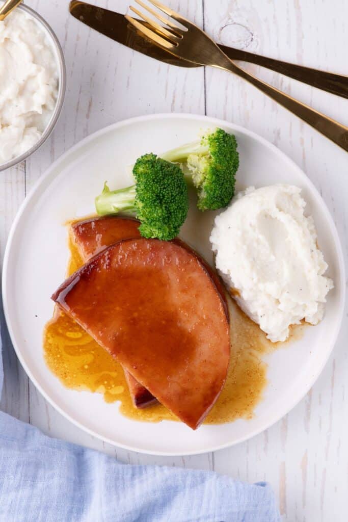 A white dinner plate with ham steak, broccoli, and mashed potatoes.