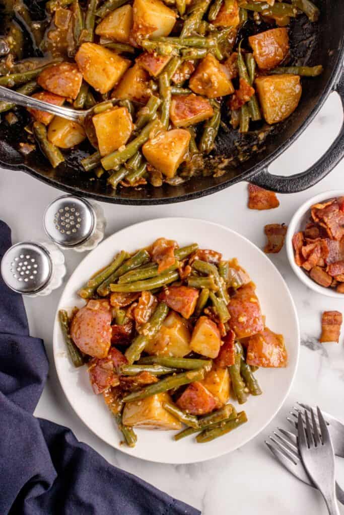 A white dinner plate with a large serving of green beans on it, next to a large cast iron pan with the smothered green beans recipe.