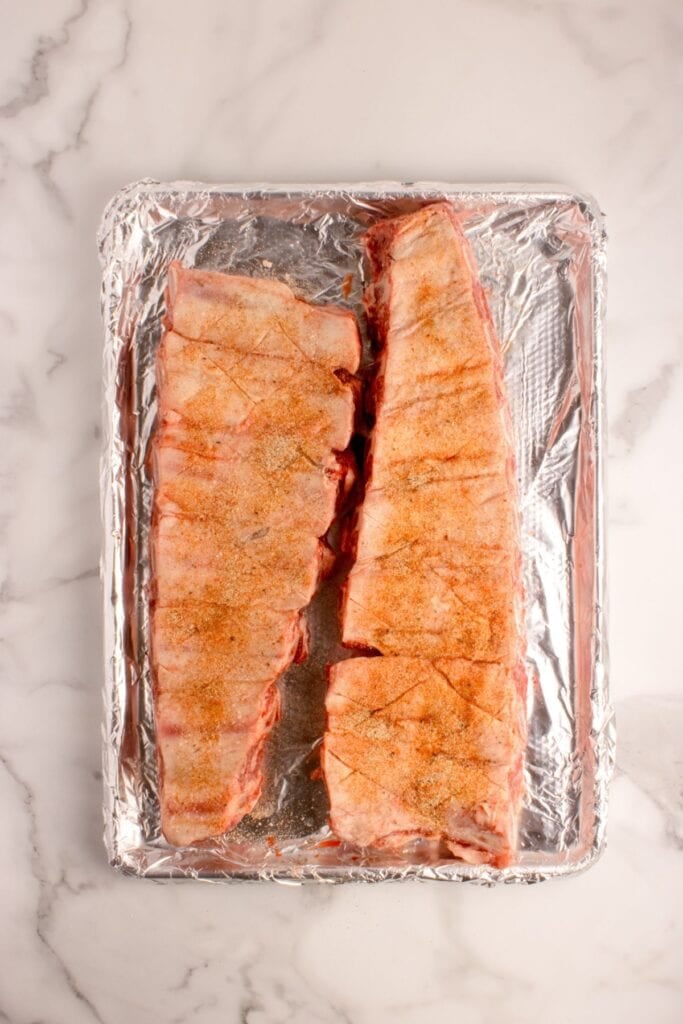 Two seasoned and uncooked racks of beef ribs on a aluminum foil lined baking sheet.