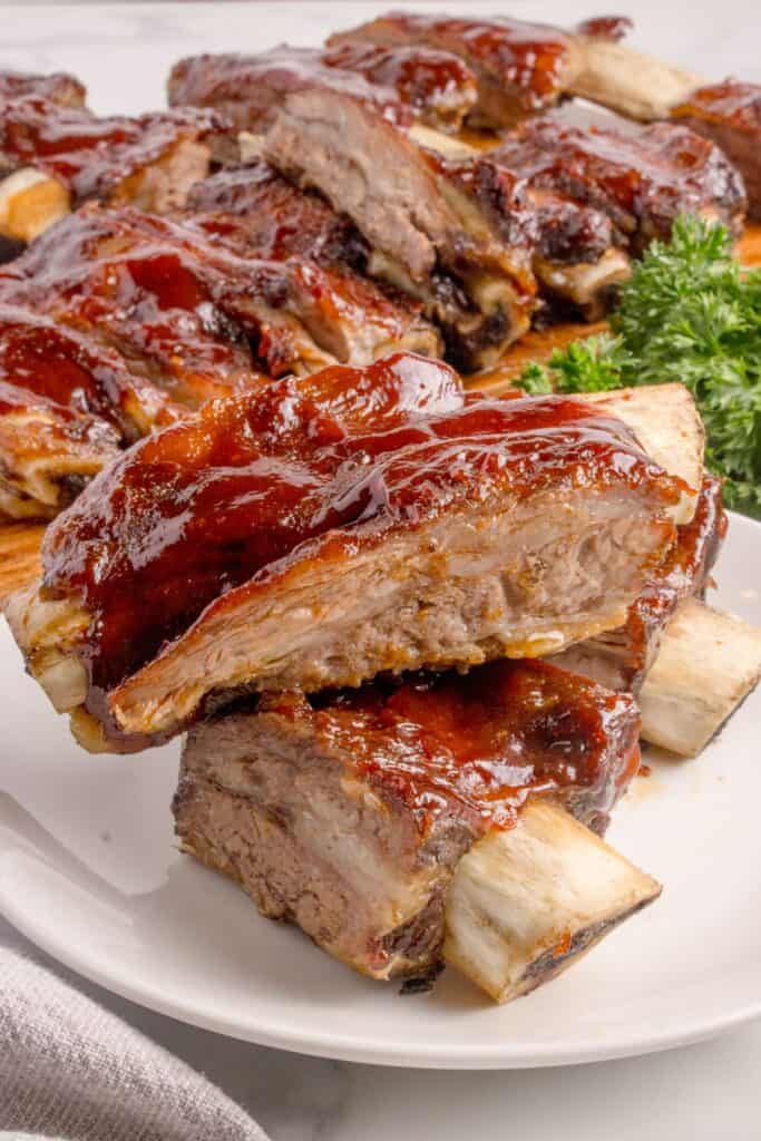 A rack of beef ribs on a wooden cutting board behind a white dinner plate with two ribs stacked on each other.
