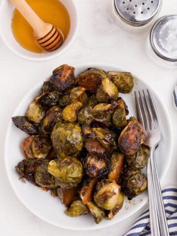 Finished brussels sprouts recipe on a white dinner plate. A small bowl of honey is beside the plate along with a pair of salt and pepper shakers.