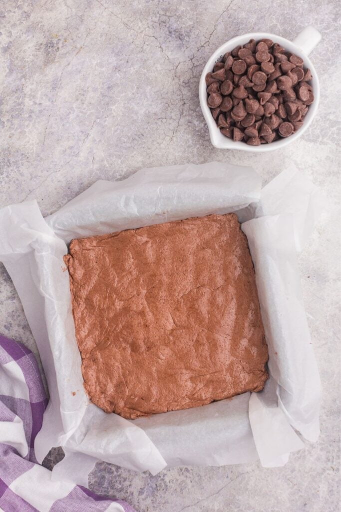 Parchment lined square baking pan with chocolate pudding and Cool Whip mixture in it. A small bowl of chocolate chips is next to the bowl.