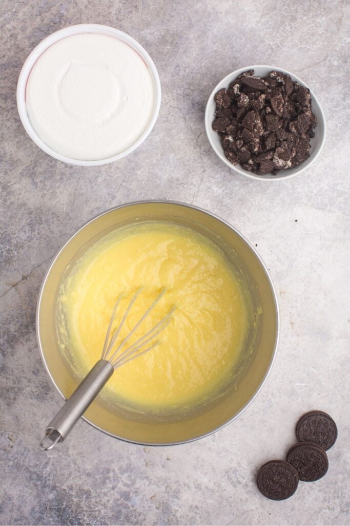 A mixing bowl with prepared vanilla pudding. A container of Cool Whip and a bowl of crushed Oreos are placed right next to the mixing bowl.