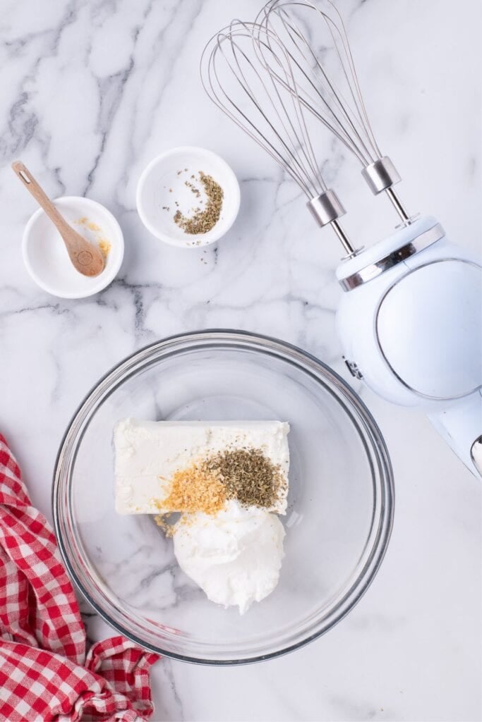 A glass mixing bowl with cream cheese, sour cream, onion, and oregano in it. A hand mixer is set beside the bowl.