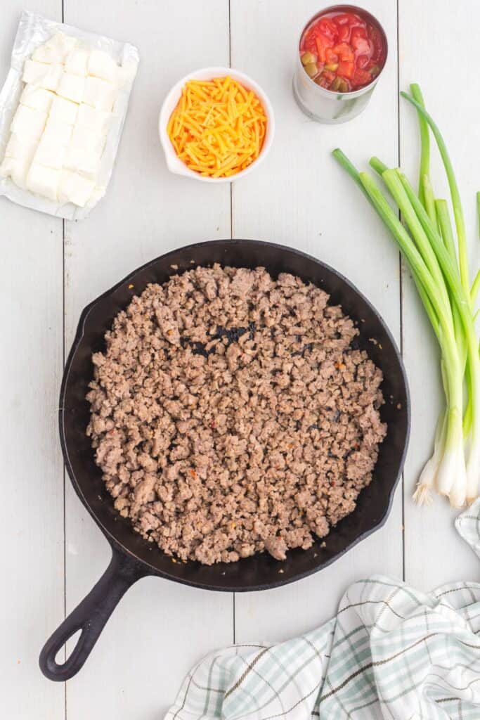 Breakfast sausage cooked and broken up in a cast iron pan. A bunch of green onions, a can of Rotel tomatoes, a cup of shredded cheese, and a package of cubed cream cheese are surrounded around the skillet.