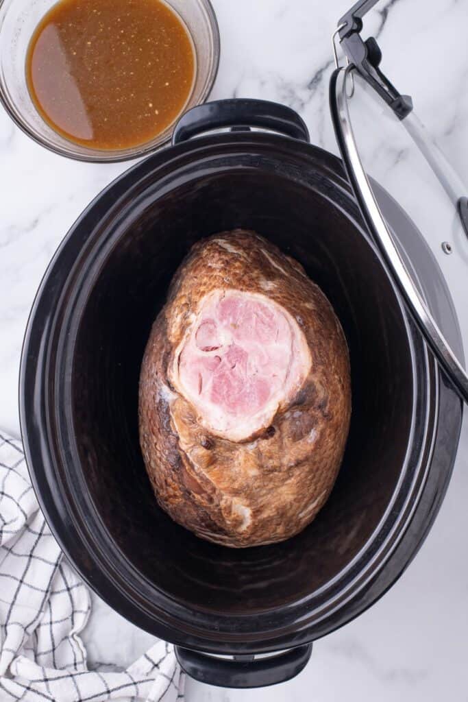 Spiral ham in a slow cooker, flat side down. A bowl of glaze is sitting next to the crock pot.