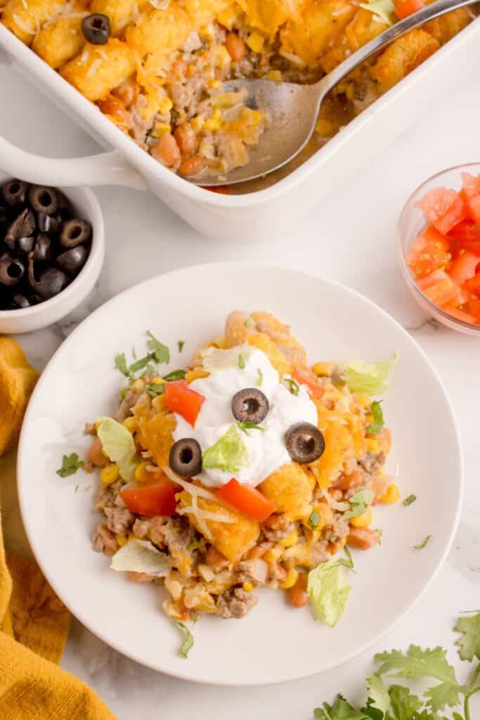 A serving of taco tater tot casserole on a white dinner plate with a dollop of sour cream, sliced olives, and diced tomatoes.