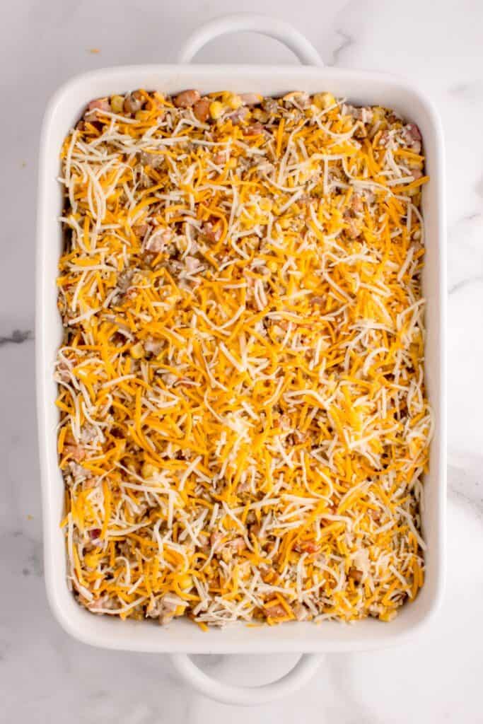White baking dish with corn, pinto bean, ground beef, onion, Rotel, and sour cream mixture at the bottom and a layer of shredded cheese on top.