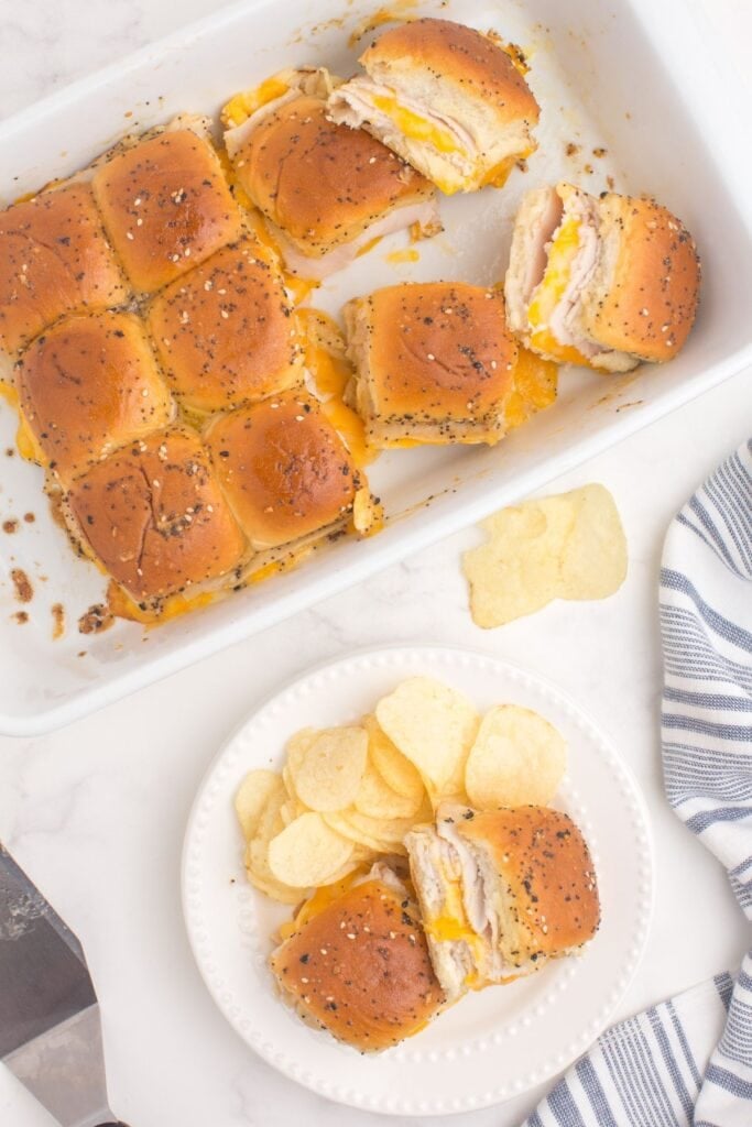 White dinner plate with two turkey sliders on it. A baking dish with the remaining turkey sliders is behind the plate. Two sandwiches are turned on their sides to show the cross section.