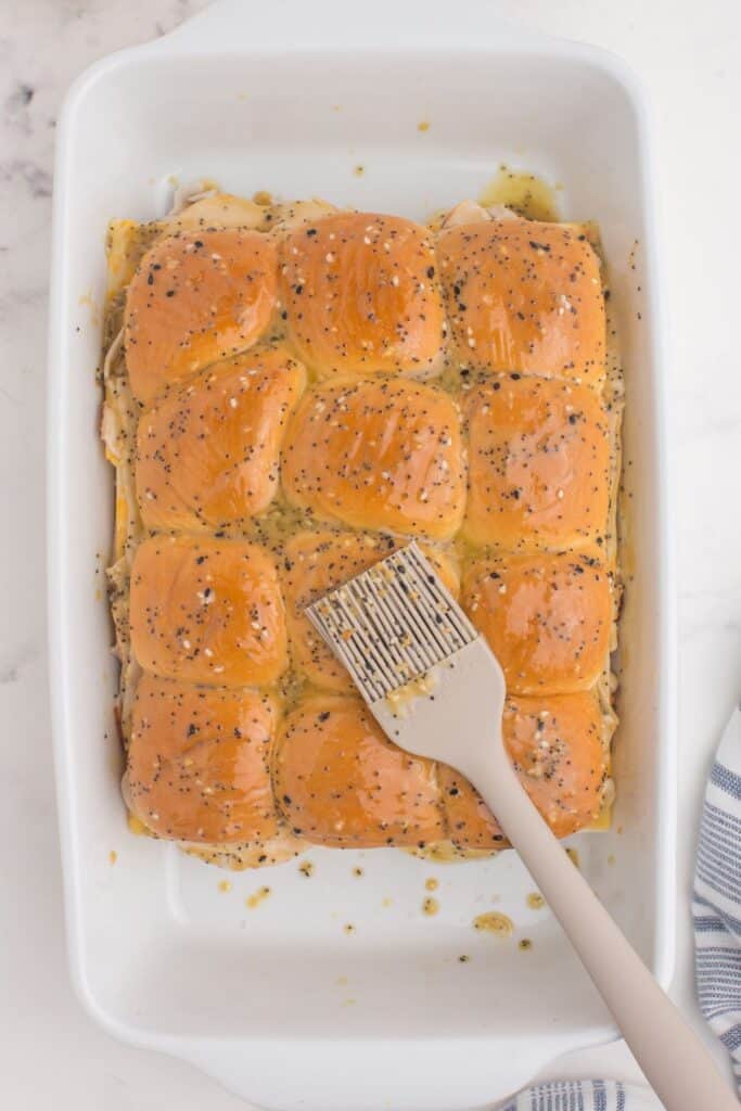 Unbaked turkey sliders in a baking dish being brushed with mixture of butter, Dijon, worcestershire, brown sugar, and everything seasoning.
