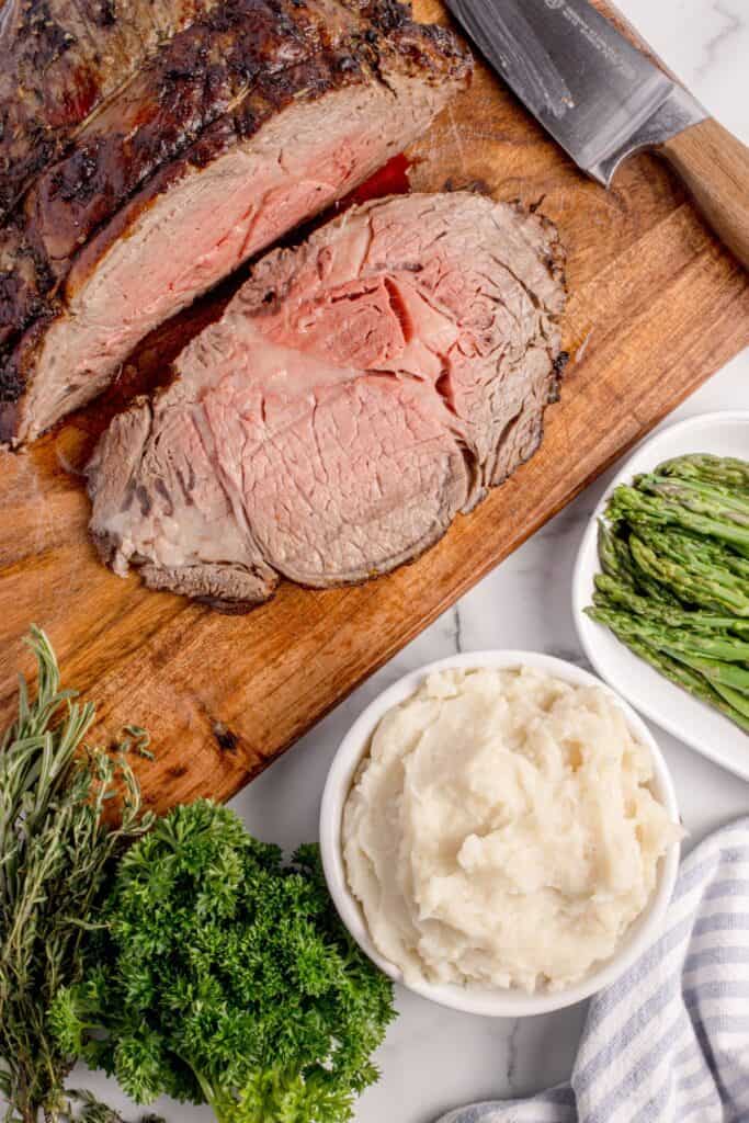 Sliced prime rib on a wooden cutting board surrounded by fresh herbs, a bowl of mashed potatoes, and a plate of asparagus.