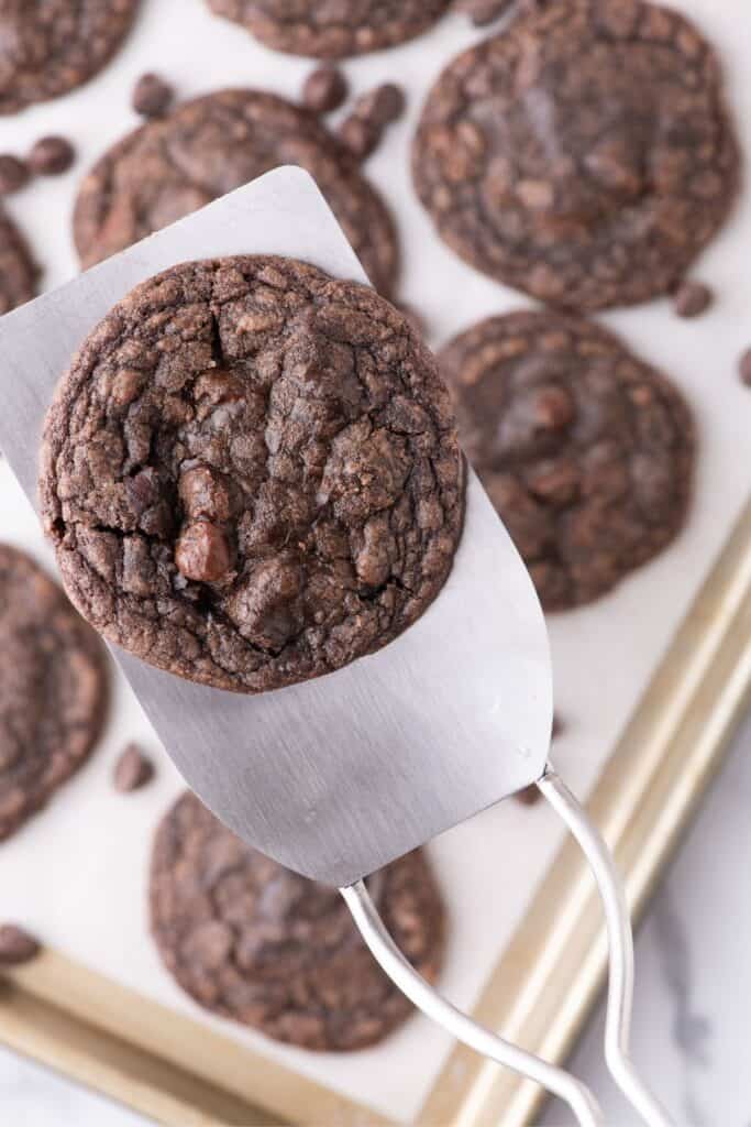 A metal spatula lifting a cookie above the baking sheet filled with baked fudge cookies.