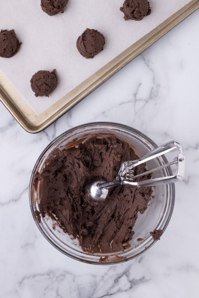 Cookie dough in a glass mixing bowl with a cookie scoop in it. A baking sheet with cookies dough balls is next to the bowl.