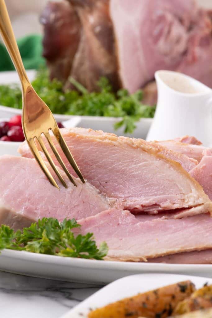 Slices of ham on a white dinner platter. A fork is lifting a serving off the plate.