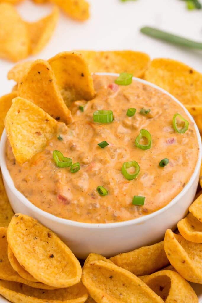 A white bowl of Rotel cheese dip with three chips lining the rim. A plate is under the bowl with corn chips surrounding the bowl.
