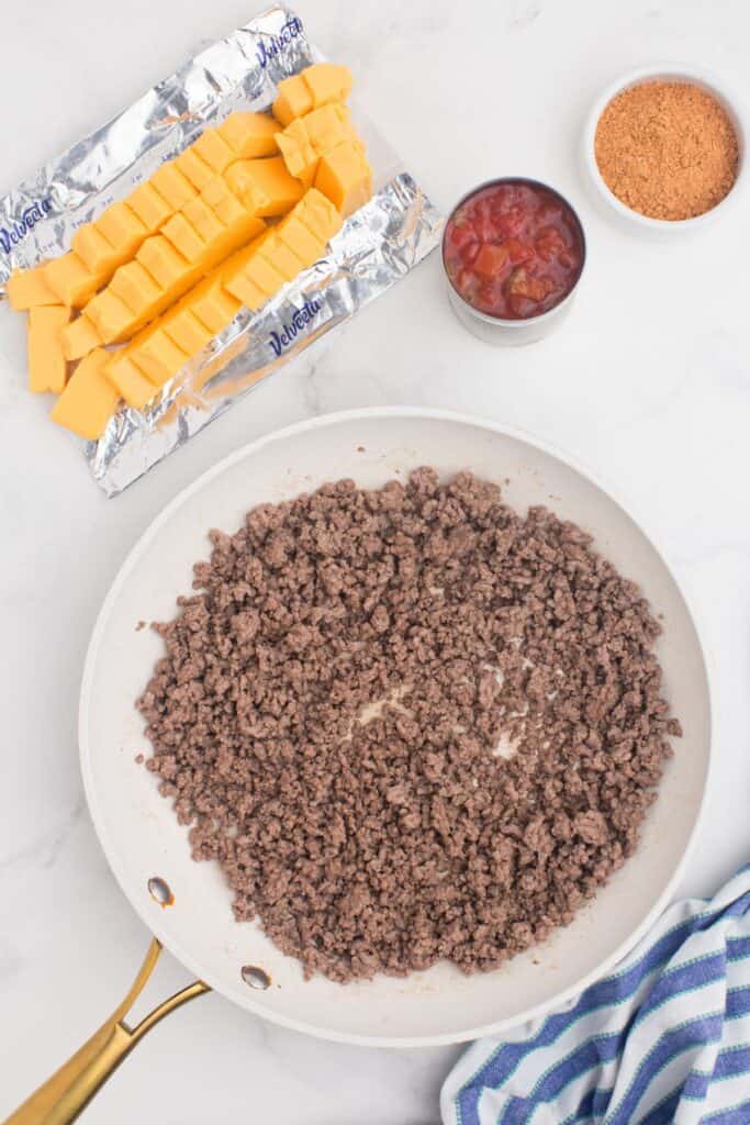 Cooked ground beef in a skillet. A can of Rotel tomatoes, small bowl of taco seasoning, and a brick of Velveeta (cut into chunks) are placed next to the skillet.
