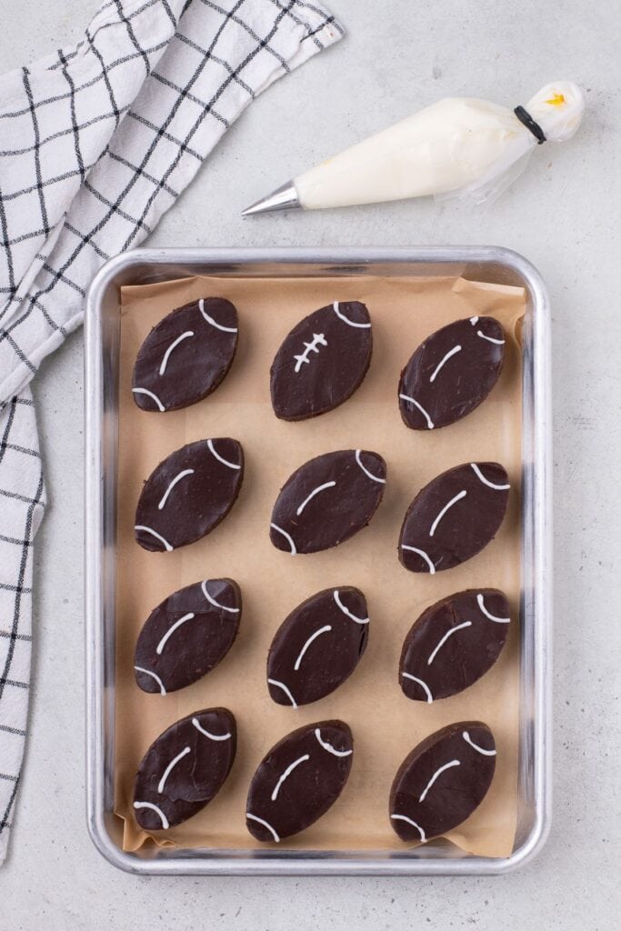 Football brownies on a parchment lined baking sheet. The icing is half completed to show how to draw the football detailing.