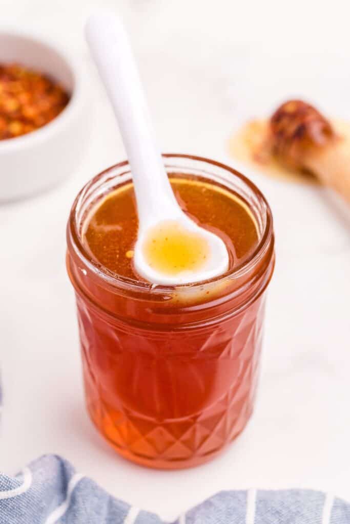 Completed hot honey sauce in a small mason jar with a white ceramic spoon resting on the surface of the sauce.