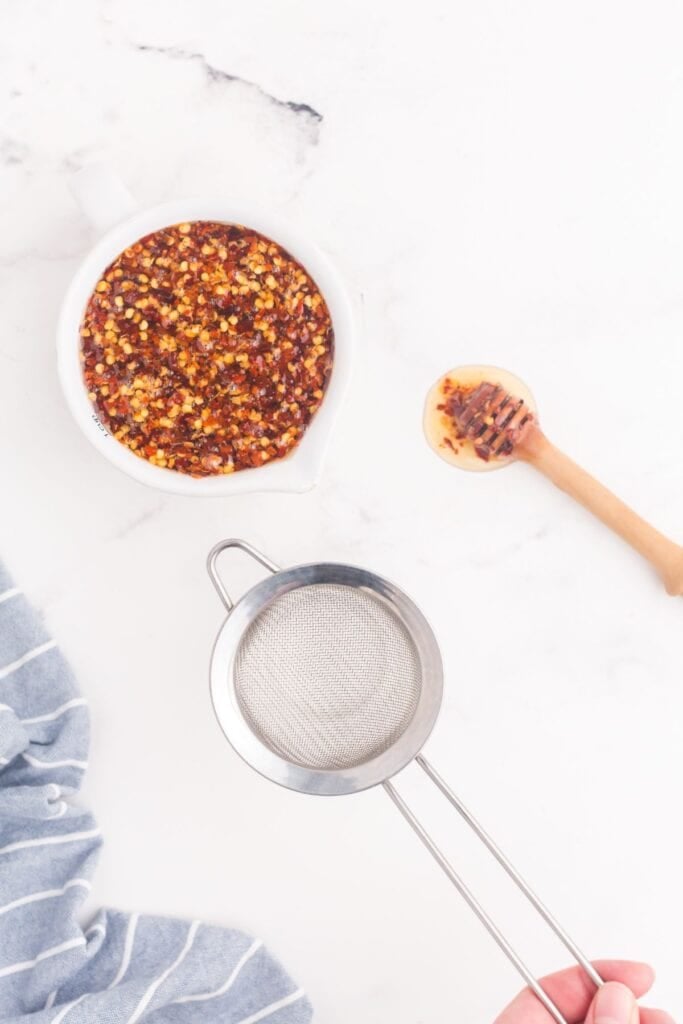 Hot honey in a pitcher next to a glass mason jar with a sieve on top of the jar.