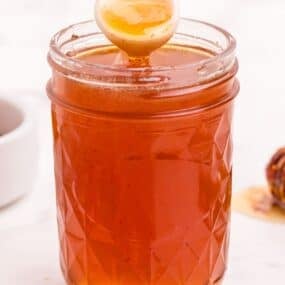 Hot honey in a small mason jar. A white ceramic spoon is lifting a scoop of honey out of the jar.
