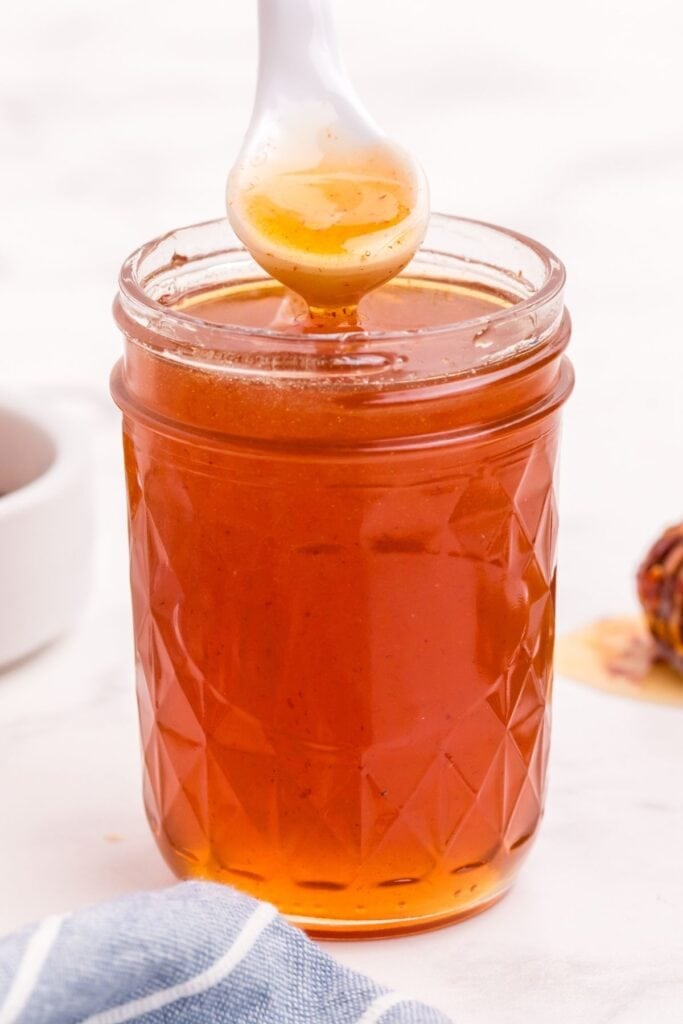 Hot honey in a small mason jar. A white ceramic spoon is lifting a scoop of honey out of the jar.