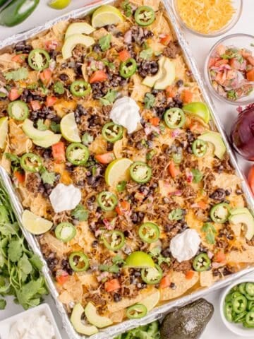 Completed oven nachos on an aluminum foil covered baking sheet surrounded by nacho toppings in bowls.