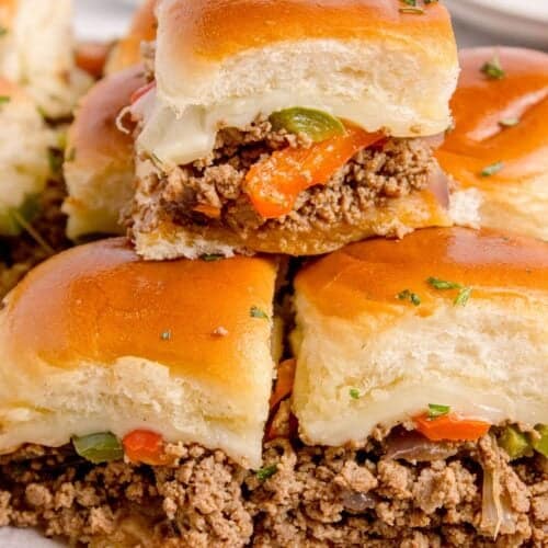 Philly cheesesteak sliders stacked on a parchment covered wooden cutting board.