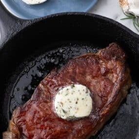 Completed compound butter on a steak in a cast iron pan. A blue plate of sliced steak butter sits above the pan.