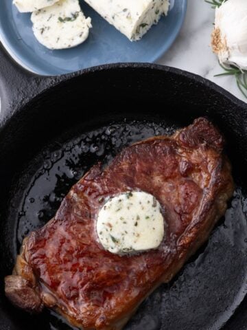 Completed compound butter on a steak in a cast iron pan. A blue plate of sliced steak butter sits above the pan.