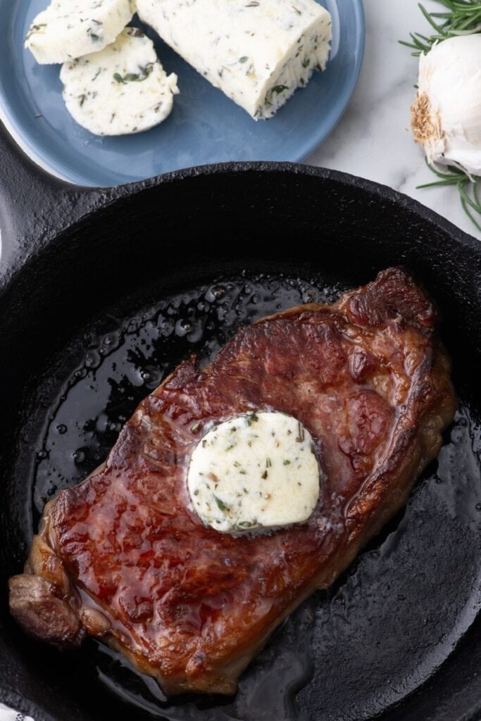 Completed compound butter on a steak in a cast iron pan. A blue plate of sliced steak butter sits above the pan.